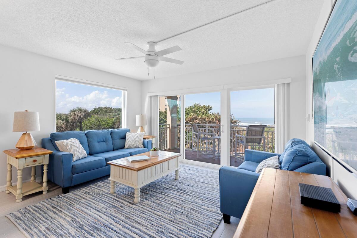 A spacious living room with a blue sofa, a wooden coffee table, and a view of the outdoors through large windows.