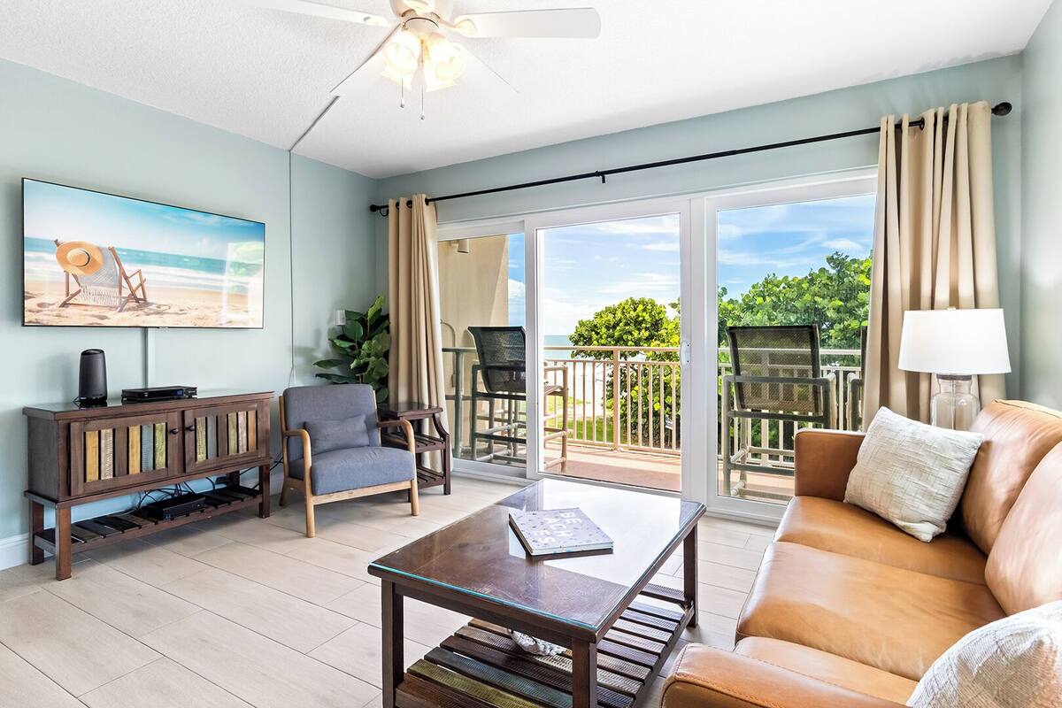 A spacious living room with a large TV, a sofa, a coffee table, and a balcony with outdoor furniture.