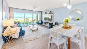 A modern living room with a large window, a dining area, and a TV. - Paradise Beach Club - Direct Oceanfront Condos with Heated Pool Pet-Friendly (Satellite Beach)