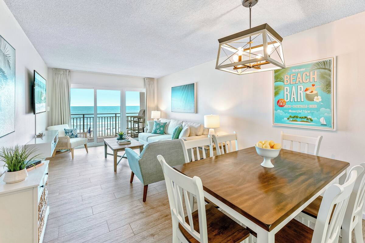 A spacious living area with a dining table, white chairs, a sofa, and a view of the ocean.