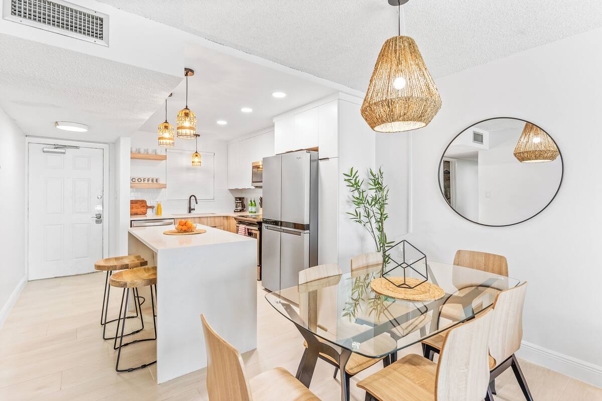 A modern kitchen with white cabinets, a central island, and a dining area with a glass table and wooden chairs.