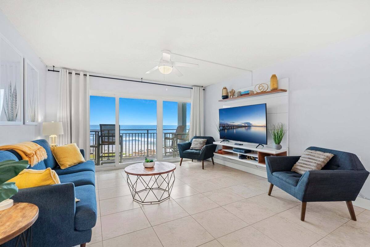 A modern living room with a large TV, comfortable seating, a balcony with a view, and a ceiling fan.