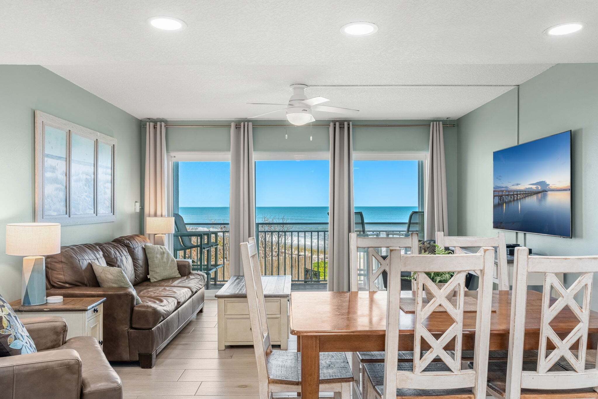 A living room with a view of the ocean, a ceiling fan, a TV, and a dining area with chairs and a table.