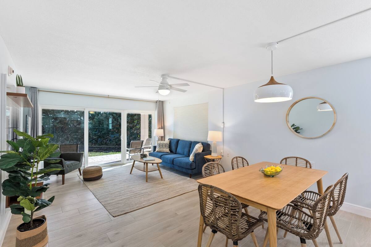 A modern living room with a wooden dining table, wicker chairs, a blue sofa, and a large window.