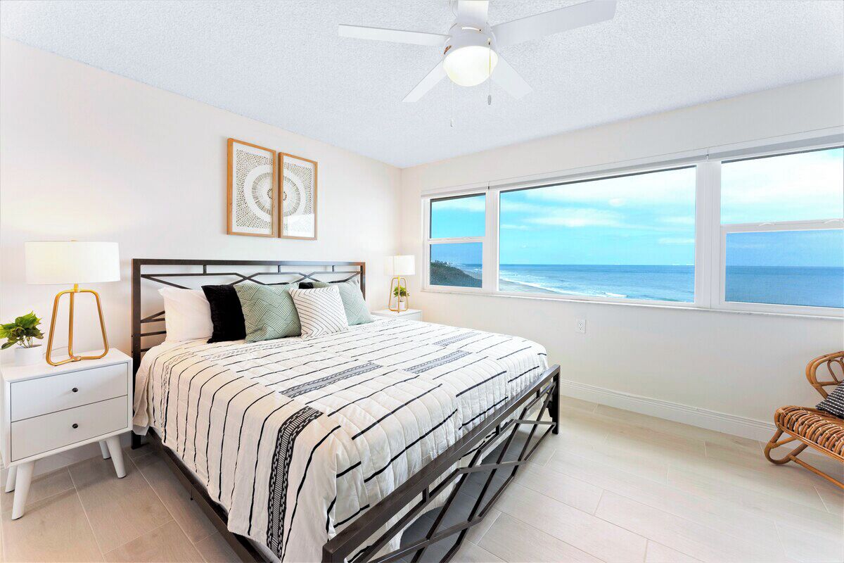 A bedroom with a bed, a nightstand, a lamp, a ceiling fan, a window with a view of the ocean, and a wicker chair.