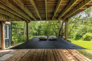 Una terraza de madera con un tapete negro, dos almohadas grandes y una estructura de pérgola de madera.