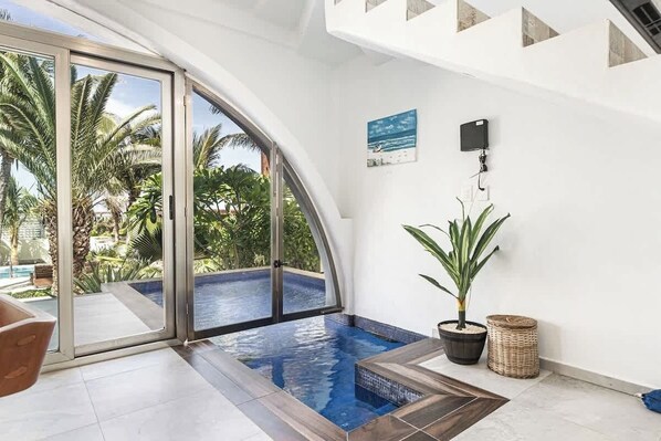 Un área de piscina interior moderna con puertas de cristal que dan a una piscina, rodeada de paredes blancas y plantas tropicales.