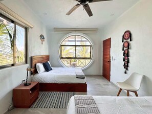 Un dormitorio con cama, mesitas de noche, una silla, ventilador de techo, ventana y puerta.