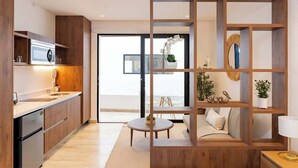 A modern kitchen with wooden cabinetry, a built-in oven, and a dining area with a round table and sofa. - Playa Studio: Rooftop Pool, Steps to 5th Avenue (Playa del Carmen)