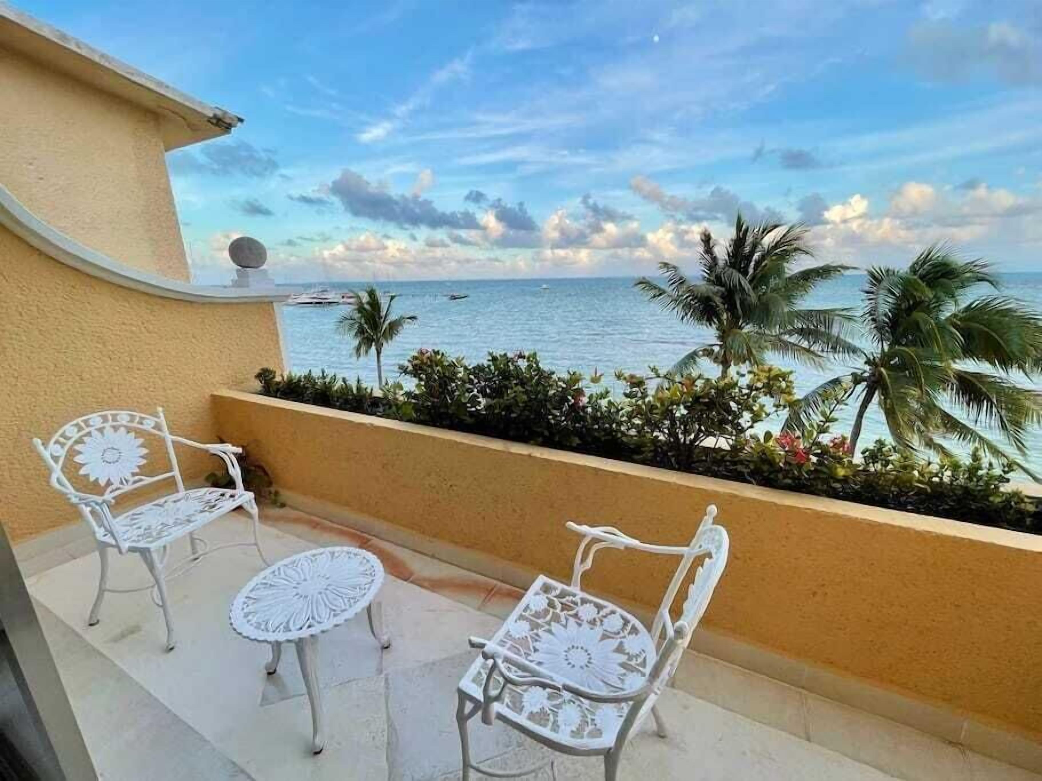 A balcony with two white metal chairs and a small round table, overlooking a beach with palm trees and boats.