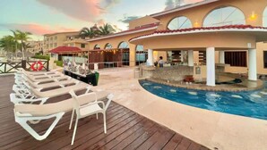 Un área de piscina en un resort con sillones de descanso, piscina y un edificio con ventanas arqueadas.