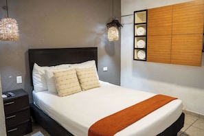 Ein ordentlich bezogenes Bett mit einer braunen Tagesdecke, ein Nachttisch mit Fernbedienung und eine an der Wand befestigte Holzkabine mit Handtüchern.