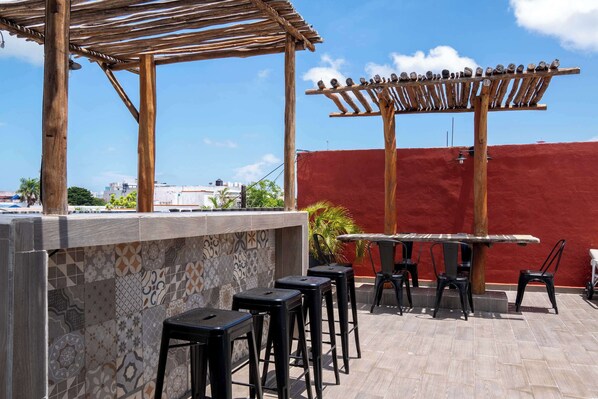 An outdoor patio with a wooden pergola, a bar area with stools, and a red wall in the background. - Central Cancun Apt - Shared Rooftop Terrace (Cancún)
