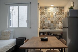 A compact kitchen with a tiled backsplash, a dining table with chairs, a refrigerator, and a window with a partially open door. - Central Cancun Apt - Shared Rooftop Terrace (Cancún)