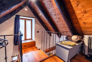 Un dormitorio de ático con techo inclinado, una ventana y una escalera de madera, y una pequeña mesita de noche con un globo terráqueo y libros.
