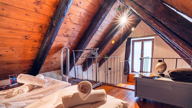 Een gezellig slaapkamer onder het dak, met houten balken, een bed met opgerolde handdoeken, een bureau met een wereldbol en een raam met gordijnen.