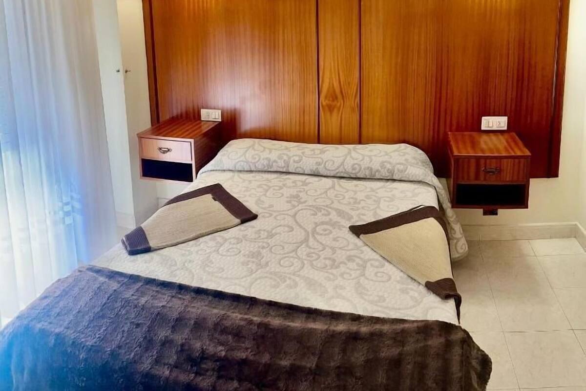 Habitación de hotel con cabecera de madera, dos mesitas de noche con lámparas, cama con ropa de cama estampada y una ventana con cortinas.