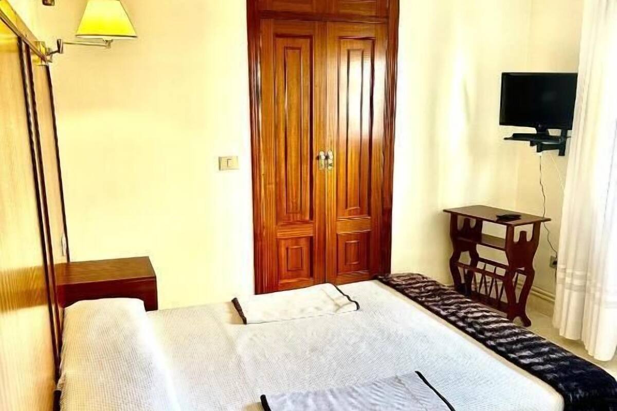 Habitación de hotel con cama, lámparas de noche, armario de madera, televisor y una mesita.