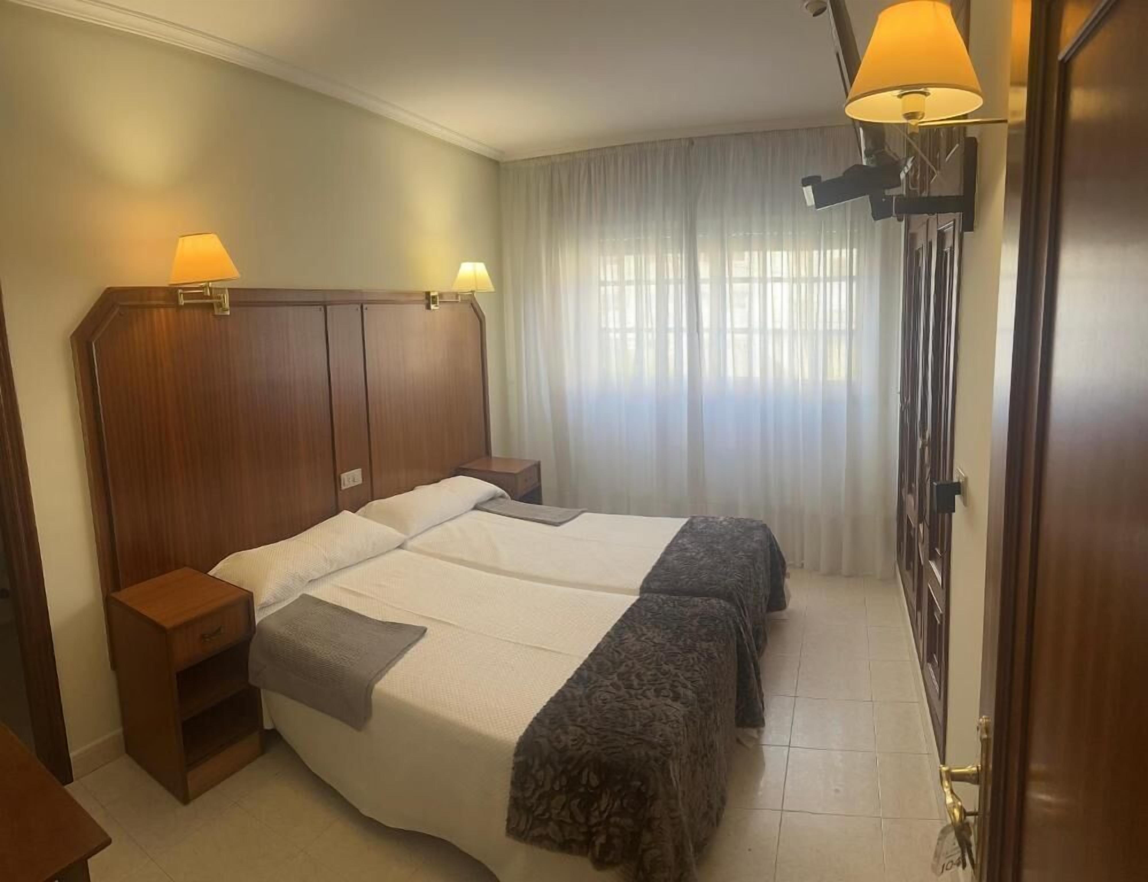 Een hotelkamer met een groot bed, twee bedlampen, een houten hoofdbord, een nachtkastje en een raam met gordijnen.