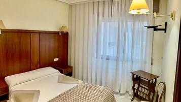 Habitación de hotel con cama, escritorio, silla, lámpara y ventana con cortinas.