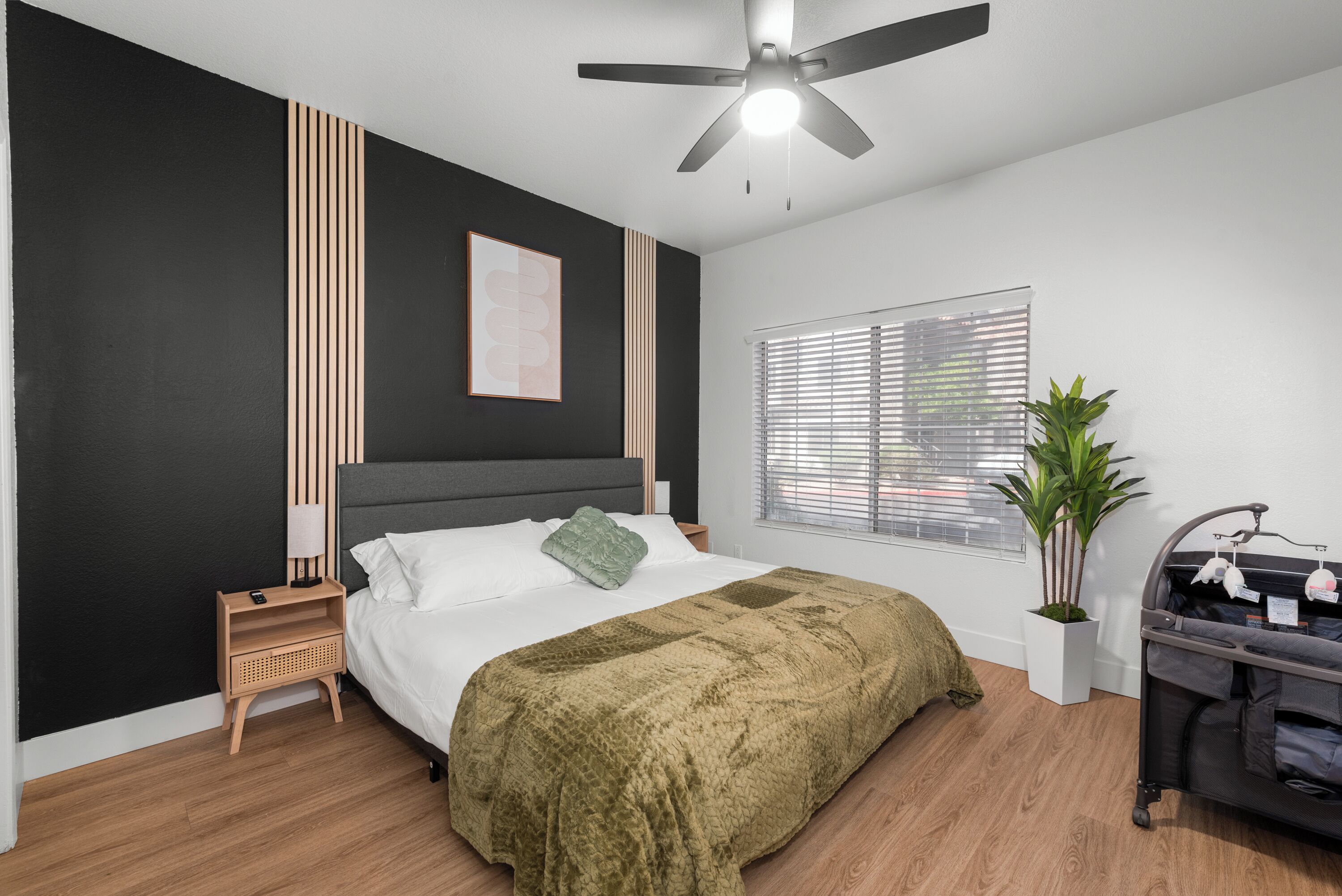 A bedroom with a bed, a ceiling fan, a bedside table, a nightstand, a window with blinds, a potted plant, and a stroller.