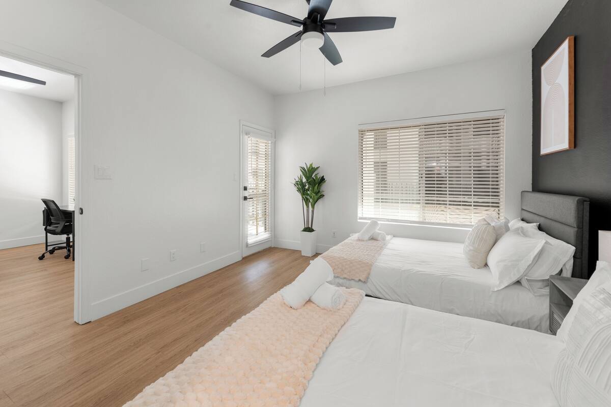 A bedroom with two beds, a ceiling fan, a potted plant, and a view of the outside through the window.