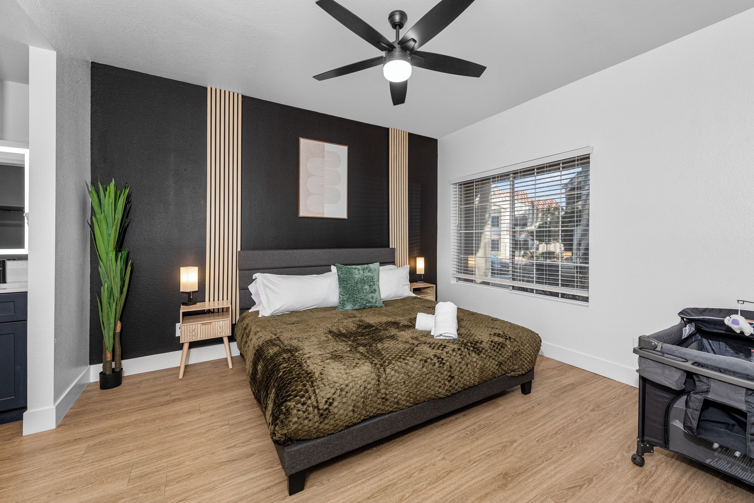 Une chambre à coucher comprenant un lit, un ventilateur de plafond, une table de chevet, une lampe, une fenêtre avec des stores et une plante.