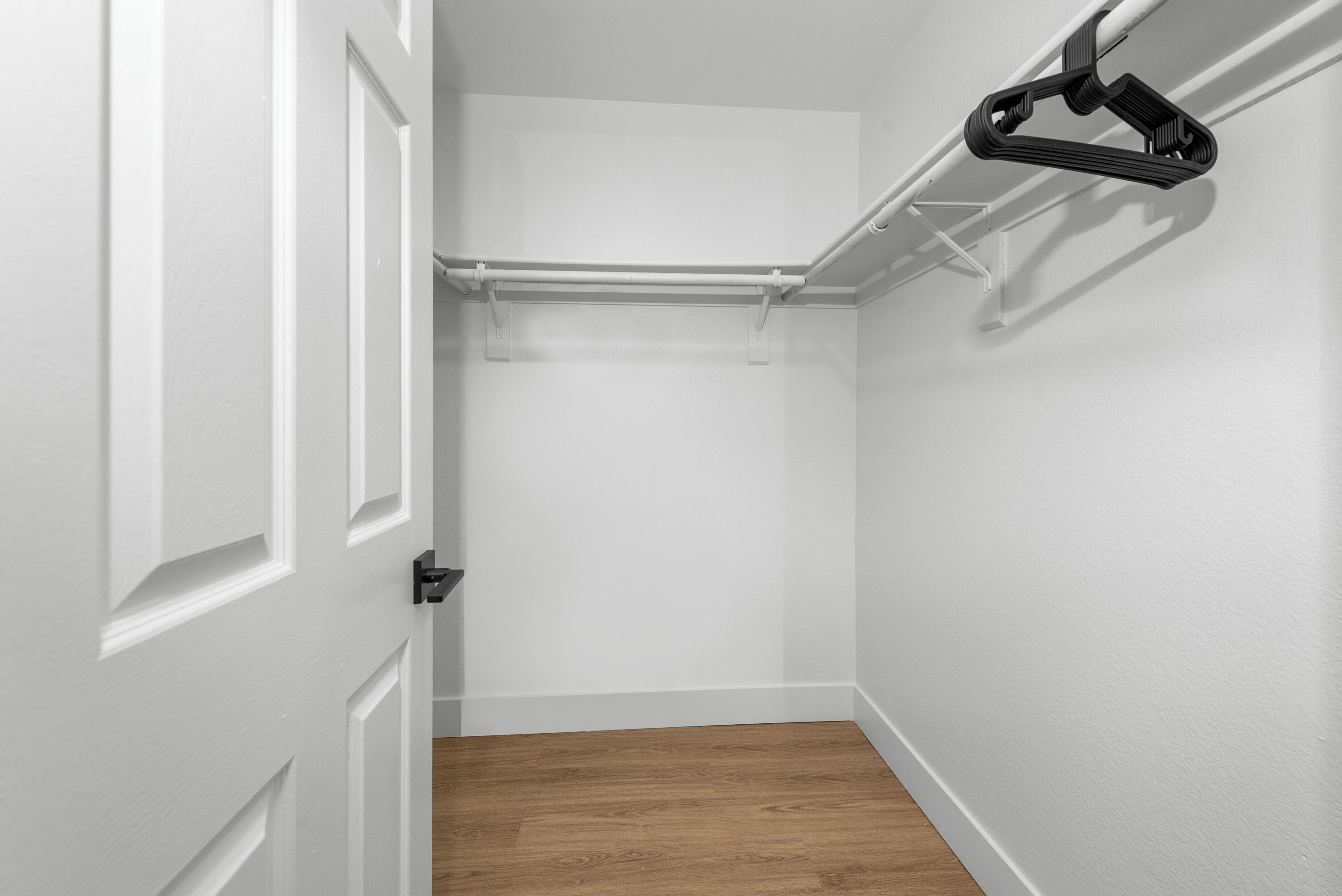Un pasillo con una puerta blanca, suelo de madera y un armario blanco con un perchero negro.