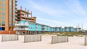 Ein Strandhotel mit mehreren Etagen, ein sandiger Strand davor und ein klarer Himmel.