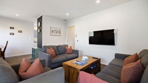 Een moderne woonkamer met een grijze zithoek, een houten salontafel en een flatscreen televisie aan de muur.