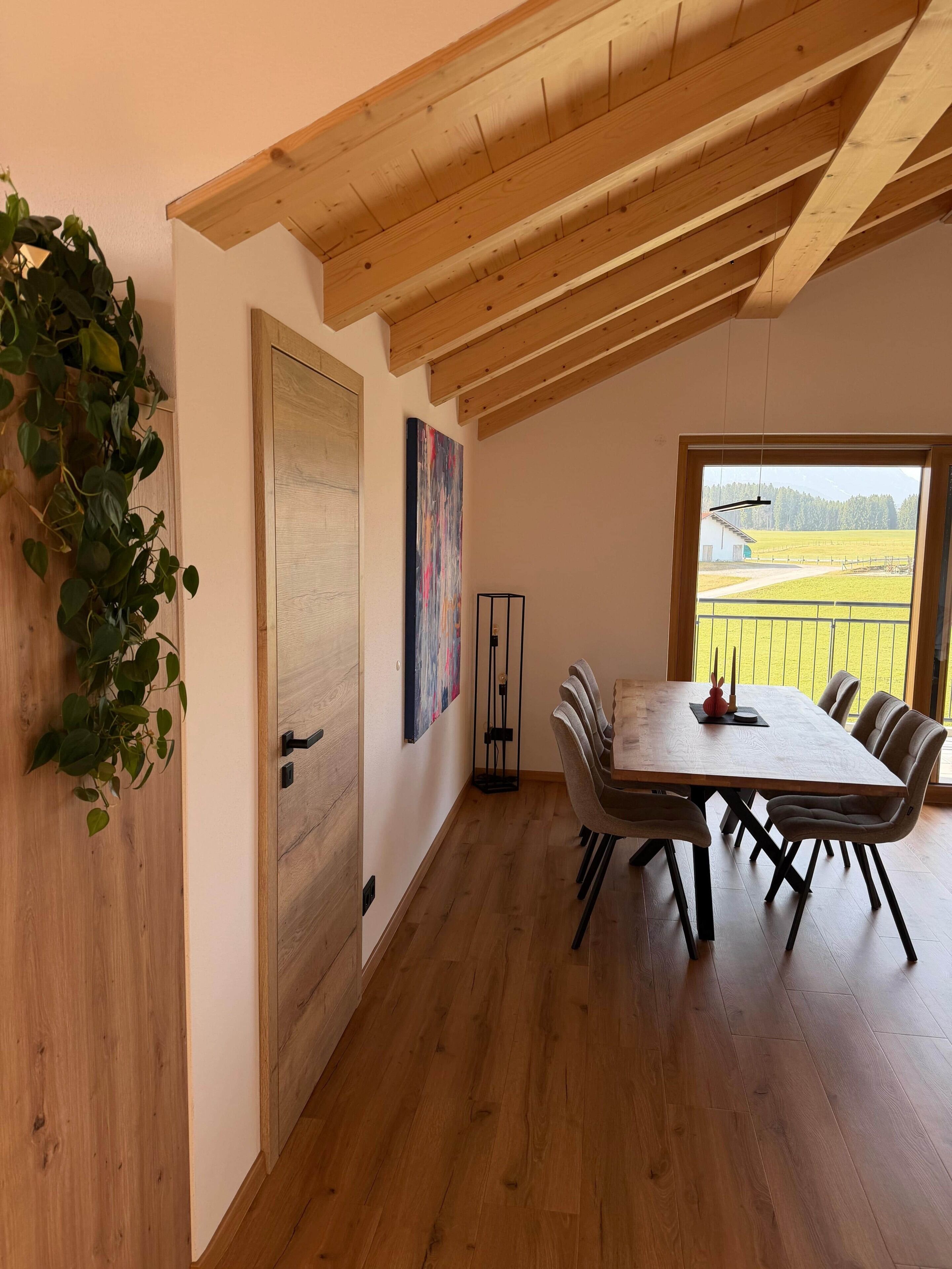 Ein Essbereich mit Holztisch und -stühlen, Blick auf ein Feld durch ein Fenster und eine Pflanze auf der linken Seite.