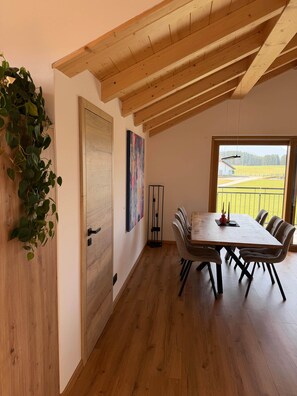 Ein Essbereich mit Holztisch und -stühlen, Blick auf ein Feld durch ein Fenster und eine Pflanze auf der linken Seite.