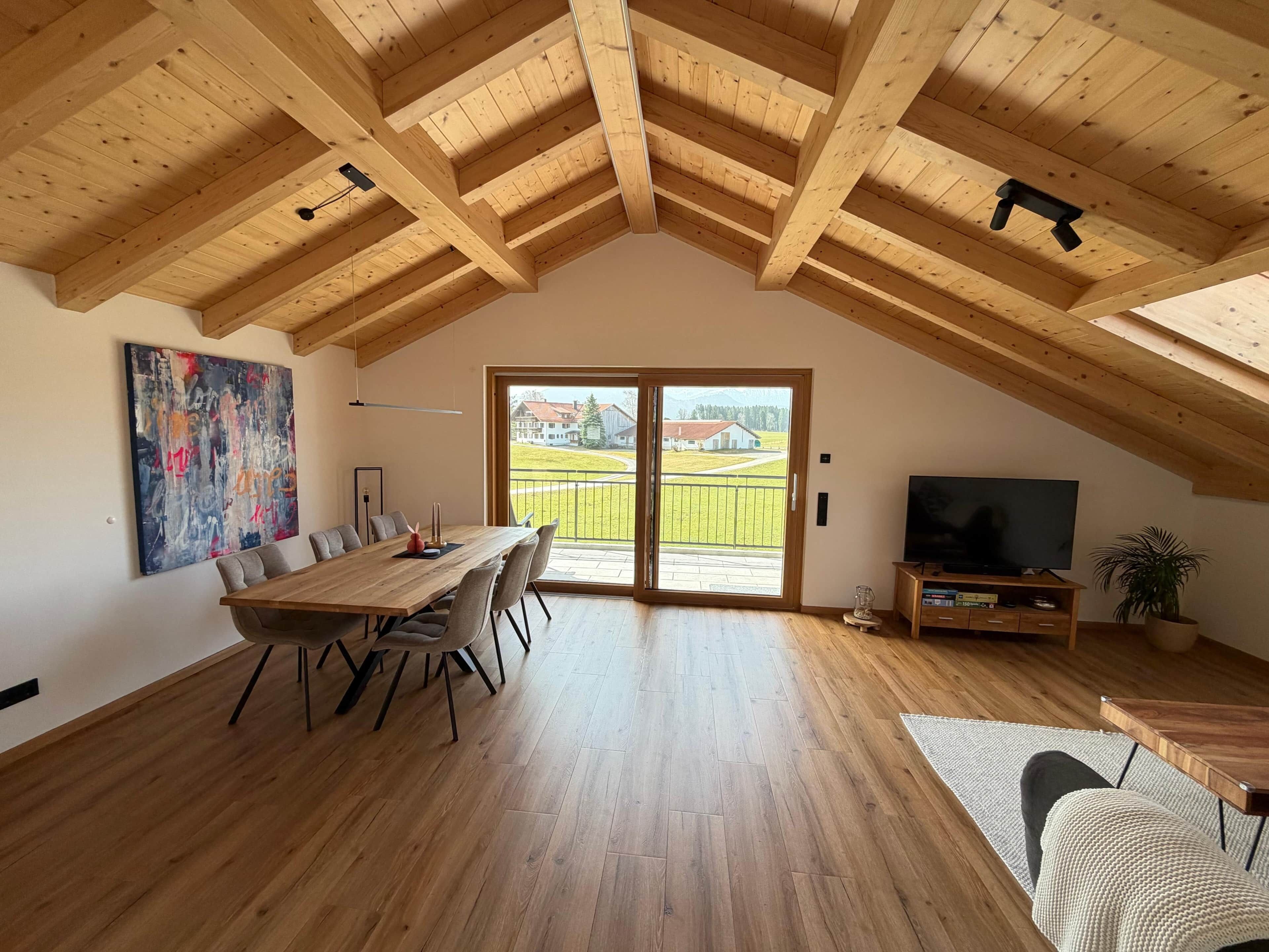 Ein geräumiges Zimmer mit hoher Holzbalkendecke, einem Essbereich mit Tisch und Stühlen, einem Fernseher und einem großen Fenster mit Blick auf eine grüne Landschaft.