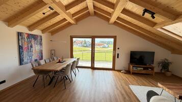 Ein geräumiges Zimmer mit hoher Holzbalkendecke, einem Essbereich mit Tisch und Stühlen, einem Fernseher und einem großen Fenster mit Blick auf eine grüne Landschaft.