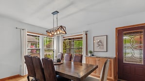 Uma sala de jantar com mesa de madeira, cadeiras de couro, luminária pendente e janelas amplas com cortinas.