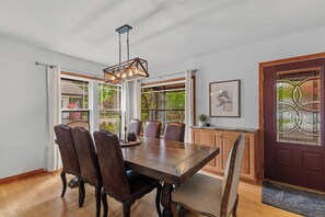 A dining room with a wooden table, leather chairs, a hanging light fixture, and large windows with curtains. - Lake City Hideaway (Coeur d'Alene)