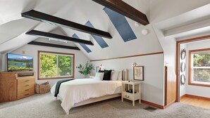 Quarto com cama, penteadeira, mesa de cabeceira, janela com vista para vegetação e claraboia.