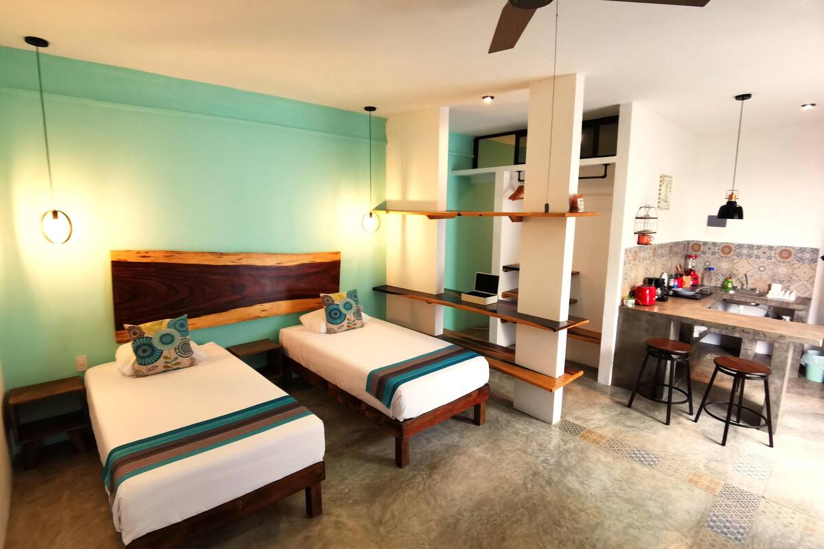A room with two beds, a kitchenette, and a ceiling fan.