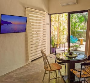 Une pièce avec un téléviseur, une porte coulissante, une table ronde avec des chaises et une vue sur la verdure à l’extérieur.