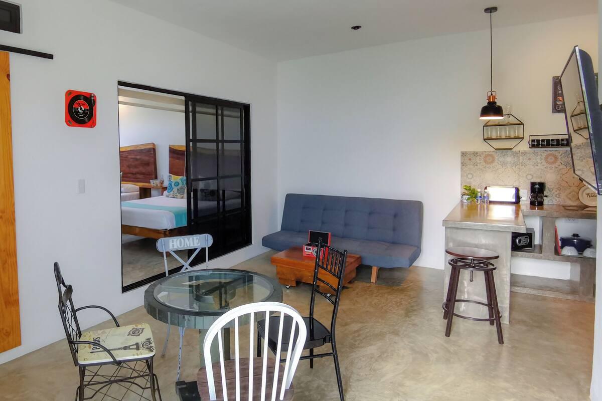 A living area with a sofa, a dining table with chairs, a kitchen area with a counter and stools, and a sliding door.