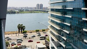 A modern high-rise hotel with a glass facade, a waterfront view, and a parking area with cars and a bus. - Luxury hotel apartment with a charming view of the Nile and the pyramids Hilton (Cairo Governorate)