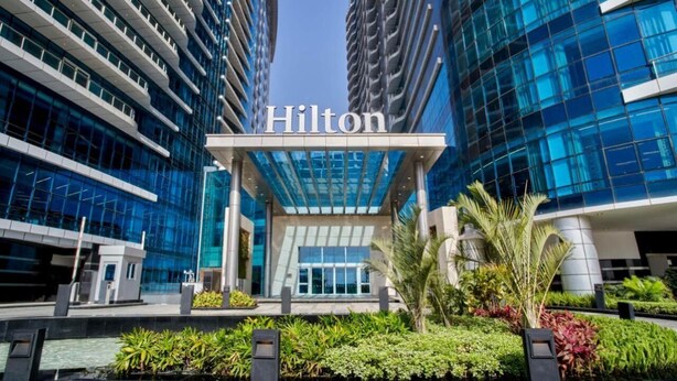 A modern Hilton hotel entrance with a glass facade, surrounded by greenery and palm trees. - Luxury hotel apartment with a charming view of the Nile and the pyramids Hilton (Cairo Governorate)