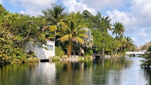 A calm river with a bridge, surrounded by lush greenery and palm trees. - Riio Shores (Miami)