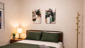 A neatly made bed with a green bedspread and pillows, a bedside lamp, abstract wall art, and a floor lamp. - Riio Shores (Miami)
