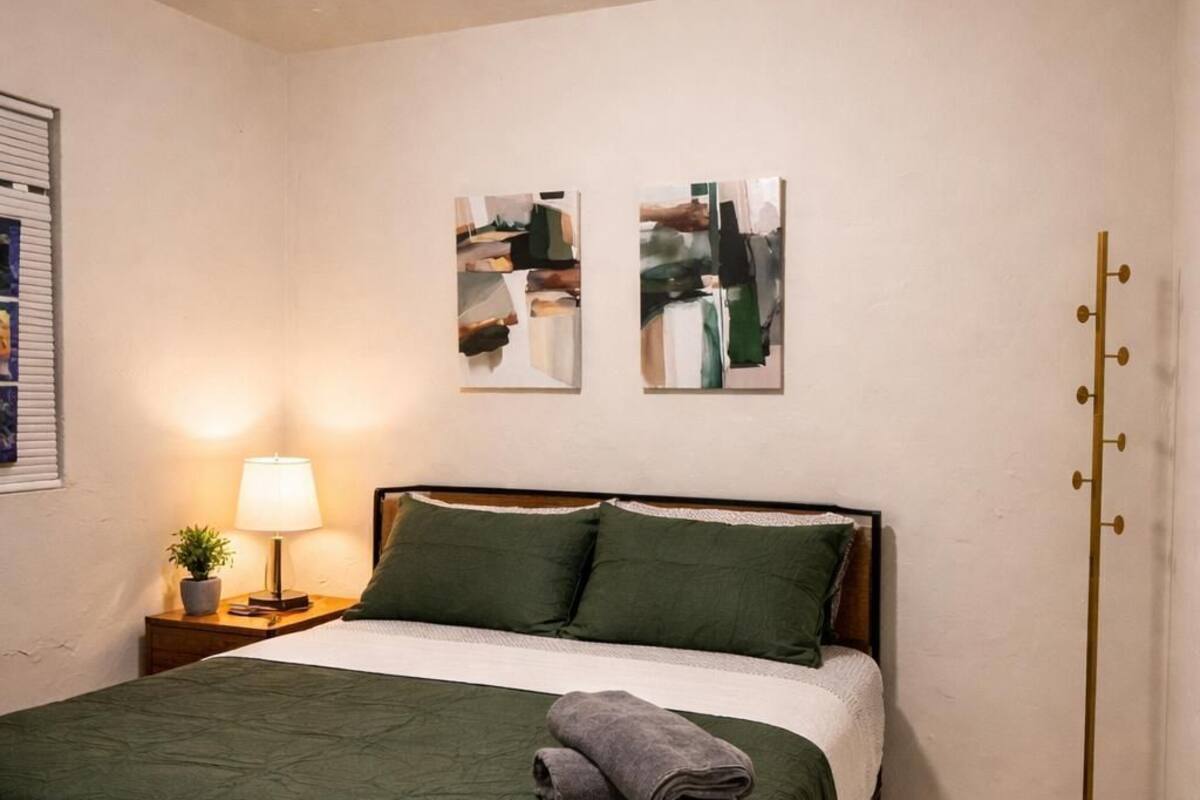 A neatly made bed with a green bedspread and pillows, a bedside lamp, abstract wall art, and a floor lamp.