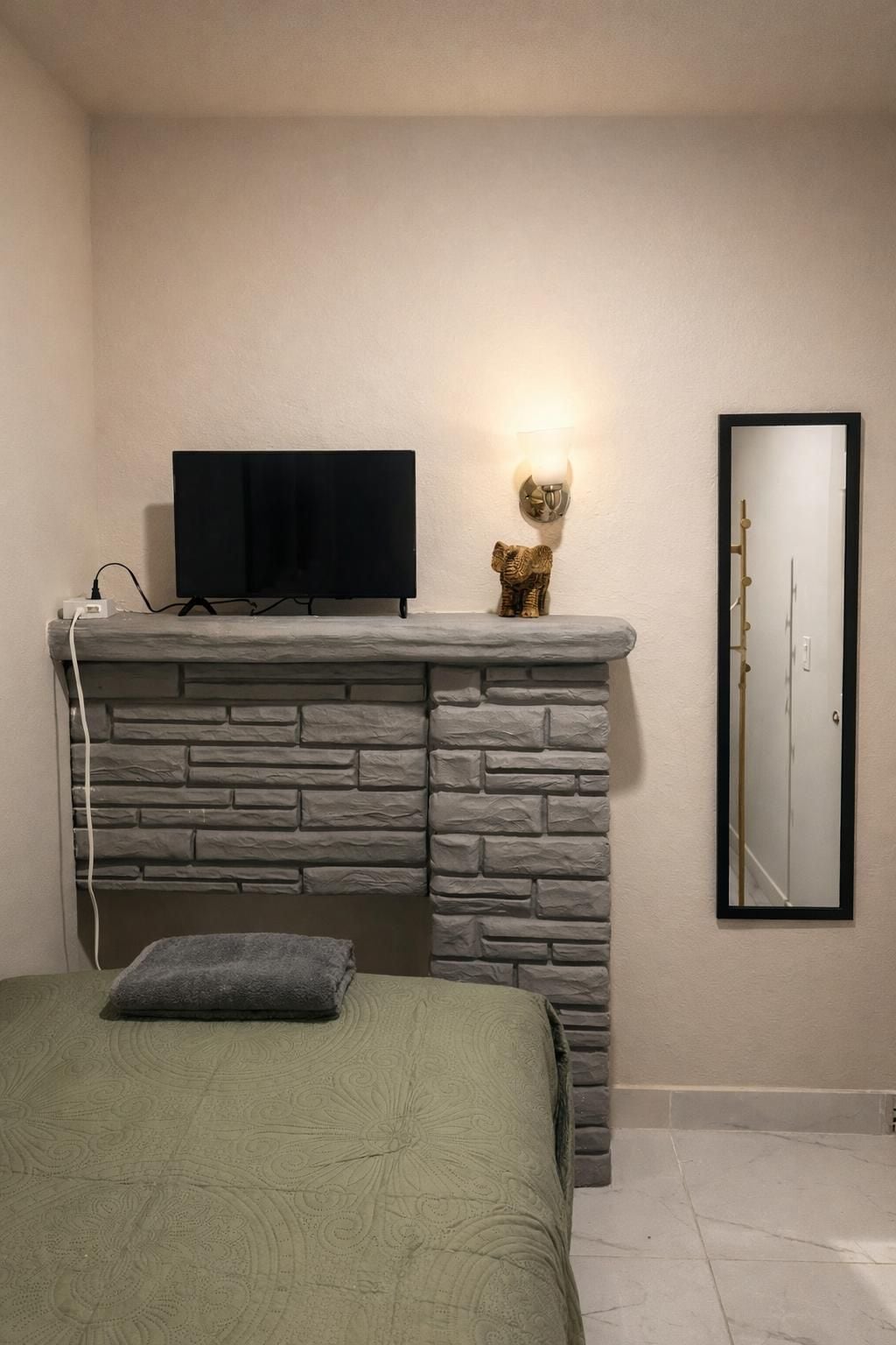 A bedroom with a stone fireplace, a television on top, a mirror on the wall, and a bed with a green bedspread.