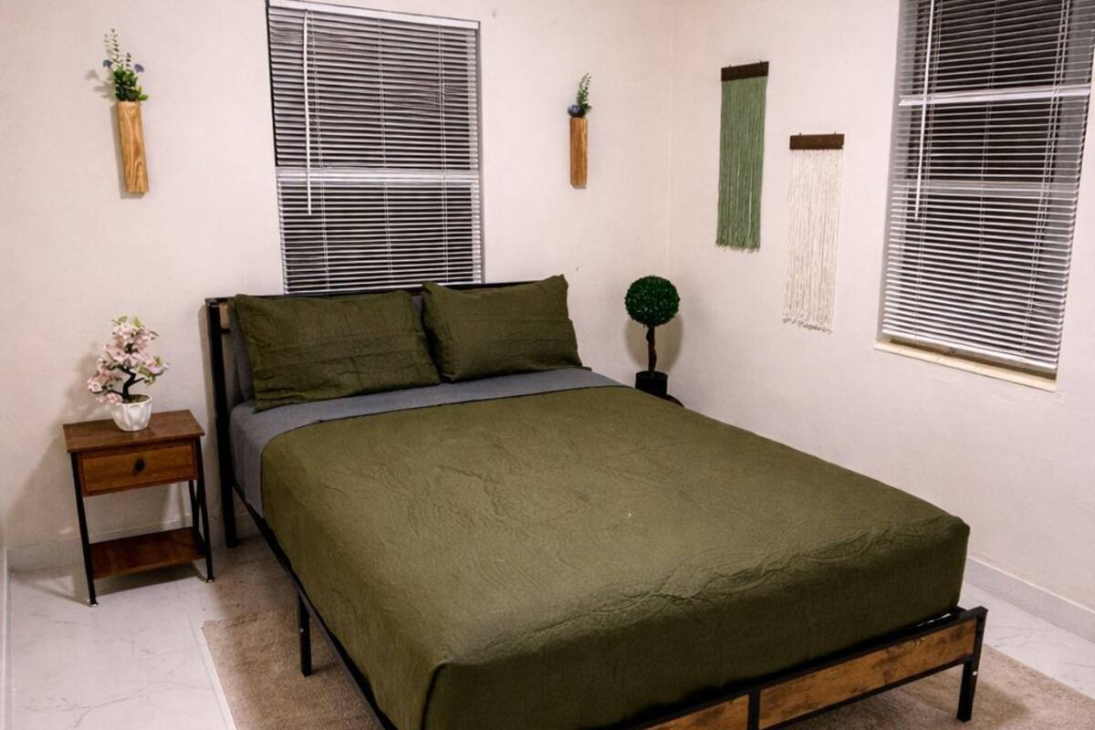 A bedroom with a bed, a nightstand, a small plant, and a window with blinds.