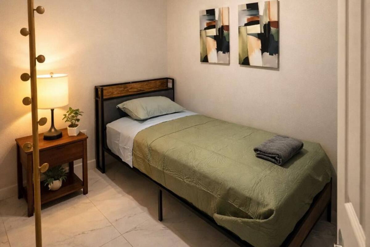 A small, neatly arranged bedroom with a single bed, a bedside lamp, a small table with a plant, and abstract wall art.