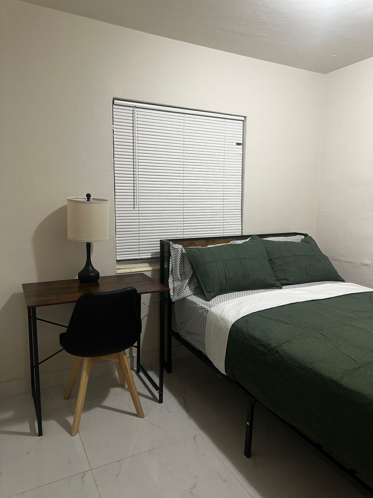 A small room with a bed, a desk, a chair, and a window with blinds.