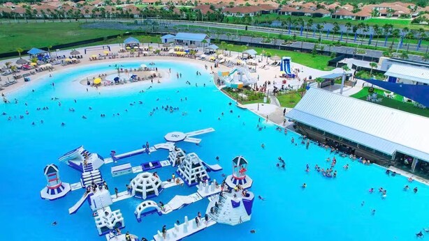 Una gran piscina al aire libre con diversas estructuras de juegos acuáticos, rodeada por una zona cubierta con asientos y un edificio.